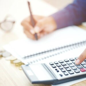 A woman writing in a notebook and using a calculator. Stacks of coins are in front of her.
